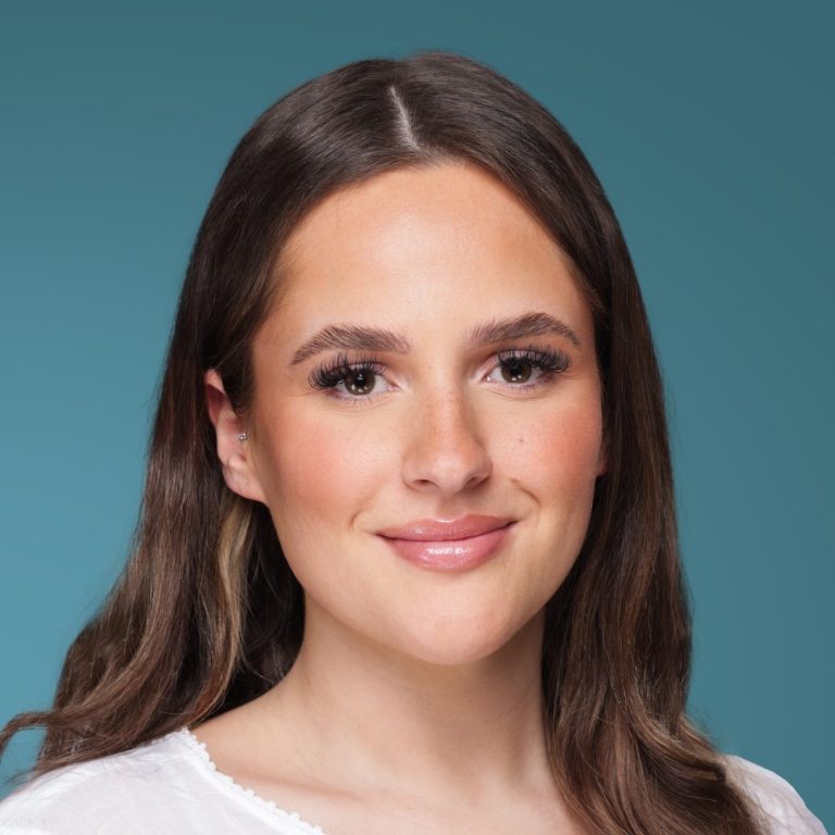 Zehra Brahimi Frau mit langen, braunen Haaren und neutralem Gesichtsausdruck vor blauem Hintergrund.