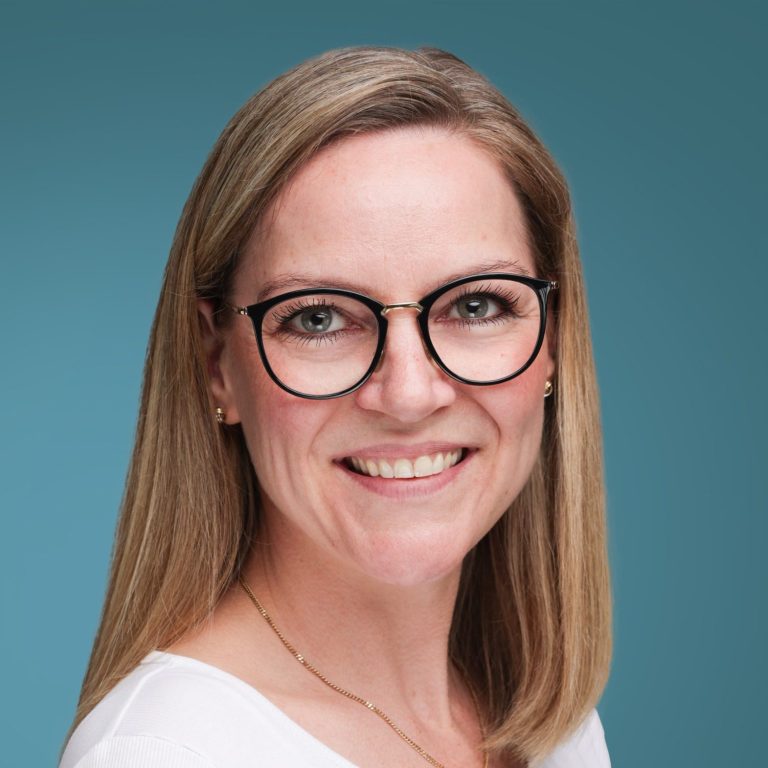 Sarah Hilger Frau mit blonden Haaren und Brille lächelt vor einem blauen Hintergrund.