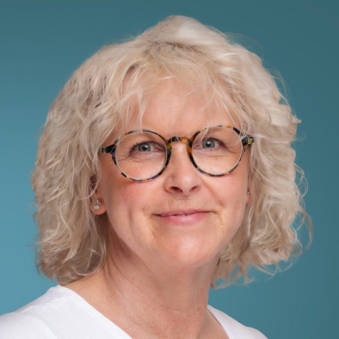 Simone Heidtmann Porträt einer lächelnden Frau mit lockigem, blondem Haar und Brille, vor blauem Hintergrund.