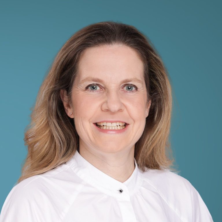 Dr. Katrin Nückel Praxisärztin Katrin Nückel, Porträt einer lächelnden Frau mit langen, blonden Haaren vor einem blauen Hintergrund.