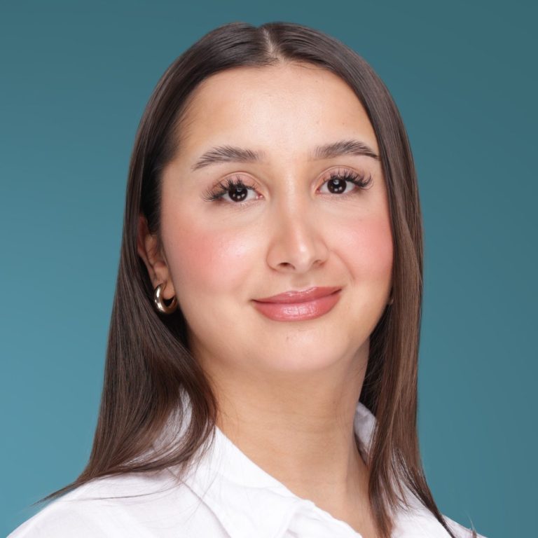 Agnesa Sahiti Lächelnde Person mit langen, braunen Haaren und weißem Hemd vor blauem Hintergrund.