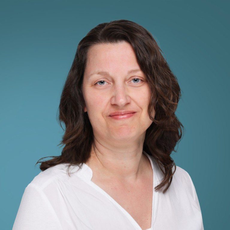 Katharina Strate Frau mit braunen Haaren, lächelnd, trägt ein weißes Oberteil vor einem blauen Hintergrund.