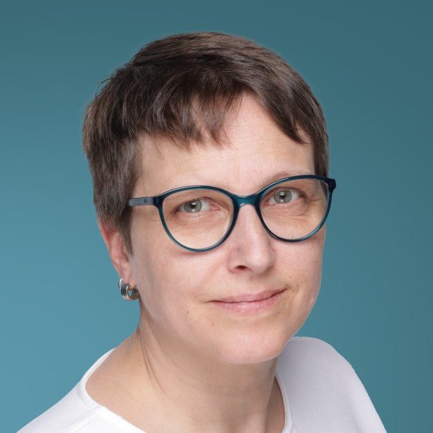 Birgit Kalkowski Frau mit Brille und kurzen Haaren, in einem weißen Oberteil, vor blauem Hintergrund.