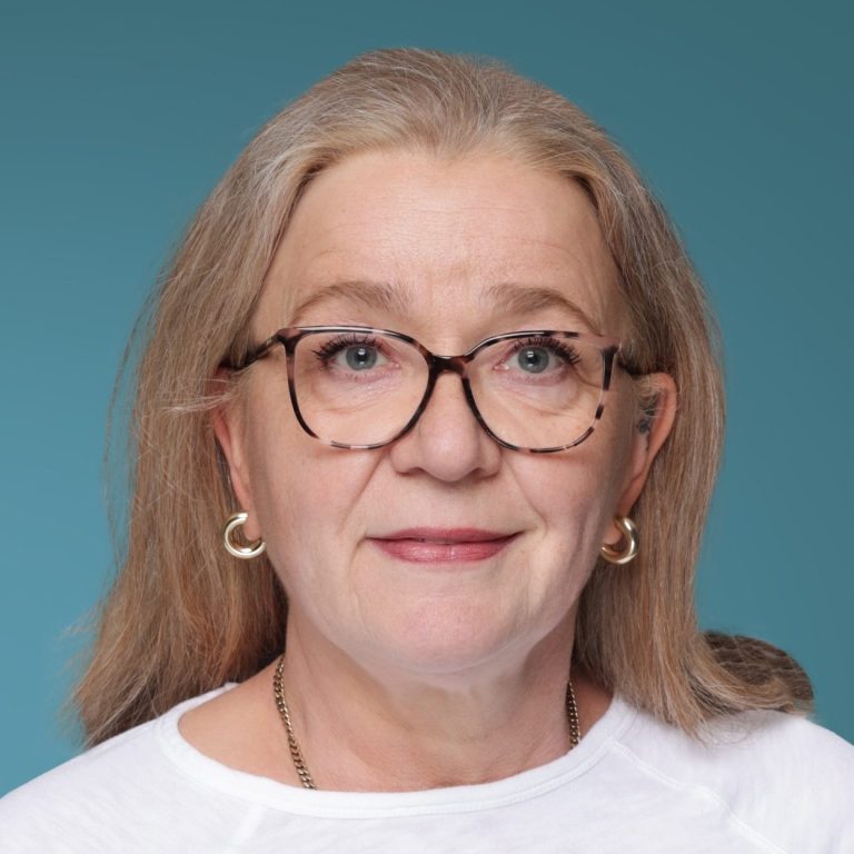 Susane Frie Frau mit Brille, blonden Haaren und einem freundlichen Ausdruck auf blauem Hintergrund.