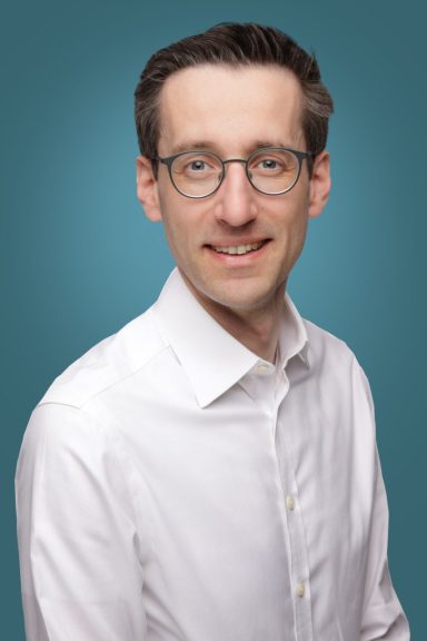 Dr. J. Matschke Porträt eines Mannes mit Brille und hellem Hemd vor einem blauen Hintergrund.